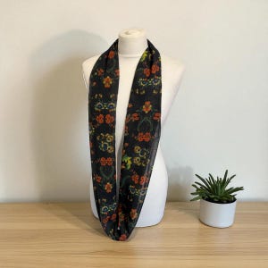 May include: A black infinity scarf with a floral pattern in shades of orange, yellow, and green. The scarf is draped on a white mannequin, showcasing its design. The scarf is made of a lightweight material.