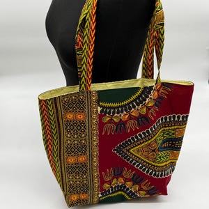 May include: A colorful tote bag with a vibrant African print. The bag features a red background with yellow, green, and black geometric patterns. The handles and trim are a mix of yellow, green, and red. The bag is displayed on a mannequin.
