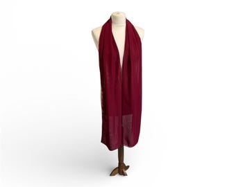 May include: A long, burgundy scarf draped over a mannequin. The scarf is made of a lightweight material and hangs down to the mannequin's waist. The scarf is a solid color with a subtle sheen. The mannequin is on a wooden stand.