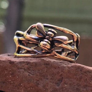 May include: A bronze-colored ring featuring a detailed bee design. The bee is centered, with wings outstretched, and surrounded by an openwork band. The ring is set against a blurred green background.