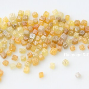 May include: A scattering of small, rough-cut diamonds in shades of yellow, orange, and brown. The gemstones are displayed on a white surface, with the text "Diamond & Craft" visible in the lower right corner.