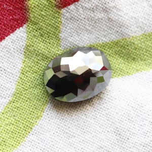 Puede incluir: Una gema gris facetada de forma ovalada. La gema refleja la luz, creando un efecto brillante. La piedra se encuentra sobre un fondo de tela blanca con rayas verdes y rojas.