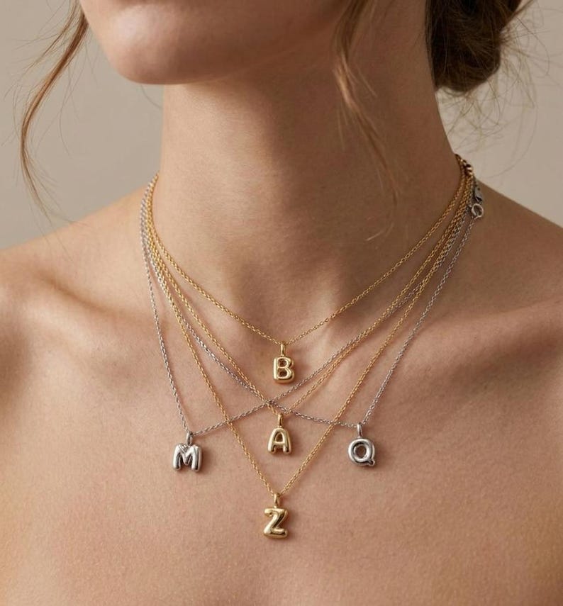 Puede incluir: Una colecci&oacute;n de collares con iniciales en oro y plata. Cada collar presenta un colgante con una sola letra, incluyendo las letras M, B, A, Z y Q. Los collares se muestran en el cuello de una persona, mostrando los diferentes estilos y colores.