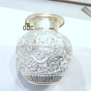 May include: A silver-colored vase with intricate floral patterns. The vase has a rounded body, a flared rim, and a detailed design covering its surface. The base has a ribbed texture. The vase is likely made of metal.
