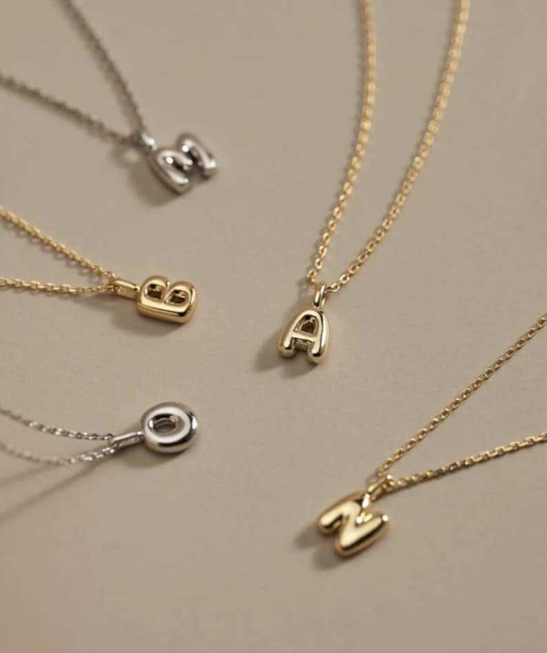Puede incluir: Una colecci&oacute;n de delicados collares con iniciales. Los colgantes tienen forma de las letras A, B, M y N. Los collares son de color oro y plata, con una fina cadena. El fondo es de color beige neutro.