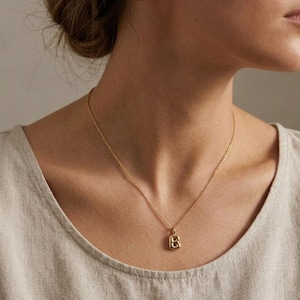 Puede incluir: Un collar dorado con una cadena fina y un colgante con la forma de la letra "B". El colgante tiene forma cuadrada con bordes redondeados. El collar se lleva sobre una parte superior de color neutro.