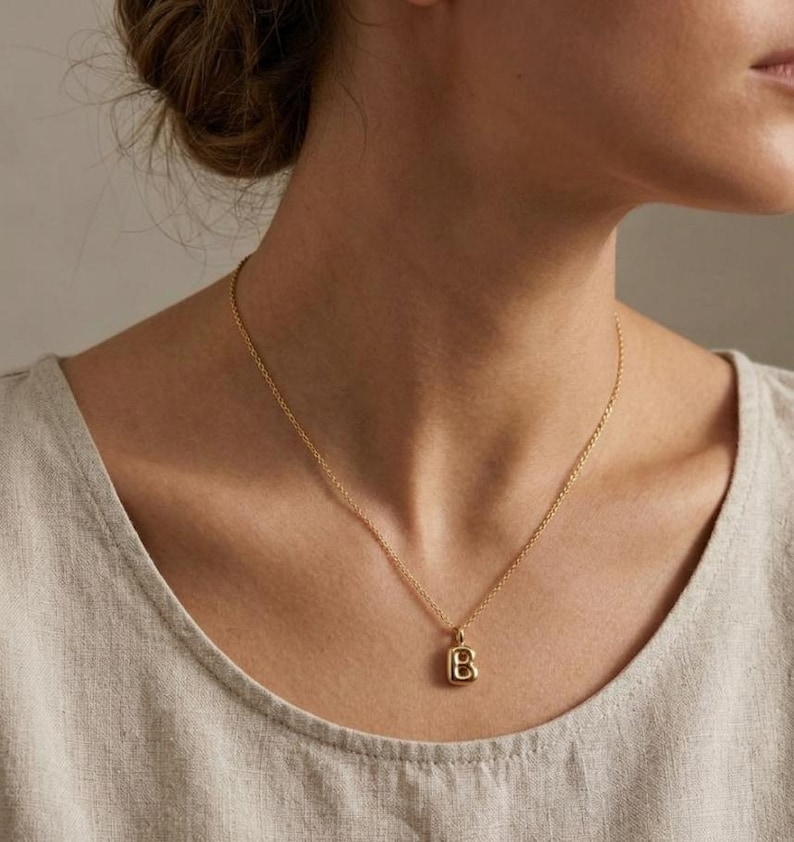 Puede incluir: Un collar dorado con una cadena fina y un colgante con la forma de la letra "B". El colgante tiene forma cuadrada con bordes redondeados. El collar se lleva sobre una parte superior de color neutro.