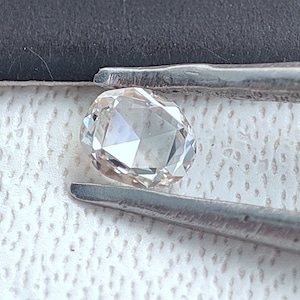 Puede incluir: Un diamante transparente de forma ovalada con un corte facetado. El diamante está sujeto por unas pinzas.