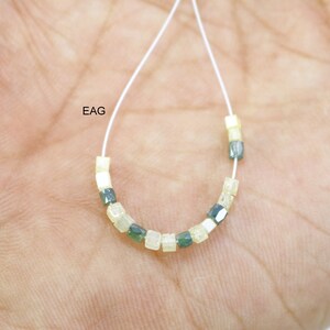 May include: A strand of beads made of alternating light yellow and dark green square-shaped beads. The beads are strung on a white thread. The text "EAG" is visible in the image.