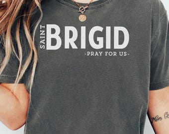 Puede incluir: Una camiseta gris con el texto "SAINT BRIGID - PRAY FOR US -" impreso en blanco.