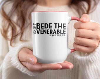 May include: A white ceramic mug with a pink interior and handle. The mug features the text "SAINT BEDE THE VENERABLE - PRAY FOR US" in black. The mug is held by a person wearing a white sweater.