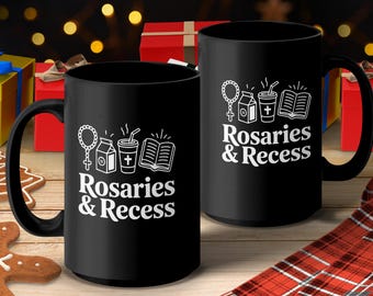 May include: Two black ceramic mugs with white text and graphics. The text reads "Rosaries & Recess" with images of a rosary, milk carton, coffee cup, and book. The mugs have black handles and are on a wooden surface.