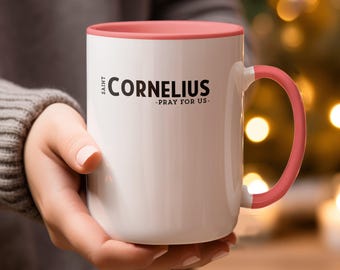 May include: White ceramic mug with a pink interior and handle. The mug features the text "Saint Cornelius - Pray For Us" in black. The mug is held by a person wearing a gray sweater.