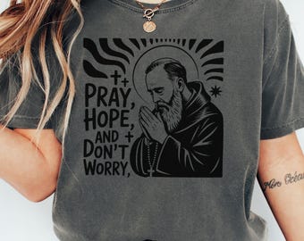 May include: Dark gray t-shirt featuring a black graphic of a praying figure with a halo, accompanied by the text "PRAY, HOPE, AND DON'T WORRY." The design includes crosses and a star. The shirt has short sleeves and a crew neck.