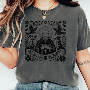 May include: Dark grey t-shirt featuring a black graphic with a religious theme. The design includes a central figure, deer, a fox, birds, and the text "LORD, MAKE ME AN INSTRUMENT".