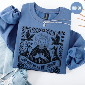 May include: Indigo blue sweatshirt with a graphic featuring a religious figure, deer, birds, and a fox. The text "LORD, MAKE ME AN INSTRUMENT" is printed on a banner. A silver necklace with a pendant is draped on the sweatshirt.