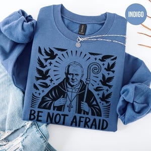 May include: Indigo blue sweatshirt featuring a black graphic of a religious figure with a staff, surrounded by doves and rays. The text "BE NOT AFRAID" is printed below the image. A silver necklace is draped across the sweatshirt.