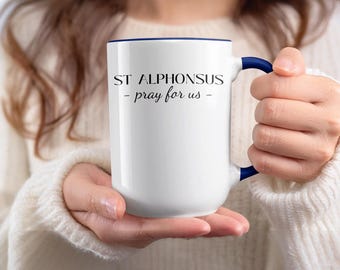 May include: White ceramic mug with a navy blue rim and handle. The mug features the text "ST ALPHONSUS - pray for us -" in black script. The mug is held by a person wearing a white sweater.