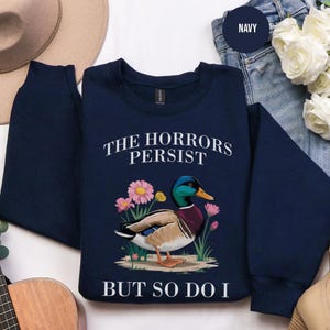 May include: Navy blue sweatshirt featuring a mallard duck illustration with pink flowers. The text reads "THE HORRORS PERSIST BUT SO DO I". The sweatshirt is shown on a neutral surface, with other items in the background.