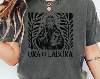 May include: Dark gray t-shirt featuring a black graphic of a religious figure holding a cross and a book. The text "ORA ET LABORA" is printed below the image, framed by decorative leaves.
