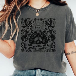 May include: Dark gray t-shirt featuring a black graphic design. The design includes a portrait of a man, doves, deer, and the text "LORD, MAKE ME A AN INSTRUMENT." The shirt has short sleeves and a crew neck.