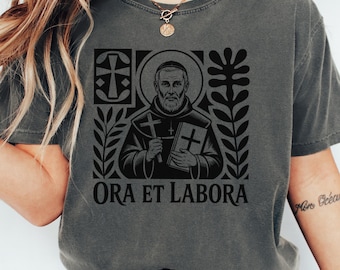 May include: Dark gray t-shirt featuring a black graphic of a religious figure holding a cross and a book, surrounded by decorative elements. The text "ORA ET LABORA" is printed below the image. The shirt has a crew neck and short sleeves.