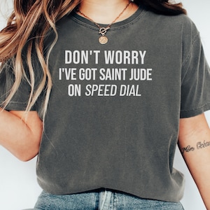 May include: Dark gray t-shirt with the text "DON'T WORRY I'VE GOT SAINT JUDE ON SPEED DIAL" in white block letters. The shirt has a crew neck and short sleeves. A gold necklace is visible.