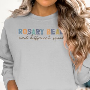 Könnte beinhalten: Ein hellgraues Sweatshirt mit dem Text "ROSARY BEAL and different speed" in einer bunten, blockartigen Schrift. Das Sweatshirt hat einen Rundhalsausschnitt und lange Ärmel. Das Thema ist Freizeitkleidung.