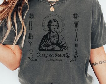 May include: Dark gray t-shirt featuring a central illustration of a woman with a halo, surrounded by sewing tools. The text "Carry on bravely - St. Zélie Martin" is printed below the image. The shirt has a vintage, worn look.