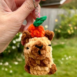 May include: A handmade keychain featuring a plush llama with a pumpkin on its head. The llama is beige and brown, with a small black face. The pumpkin is orange with green leaves. The keychain is attached to a silver ring.
