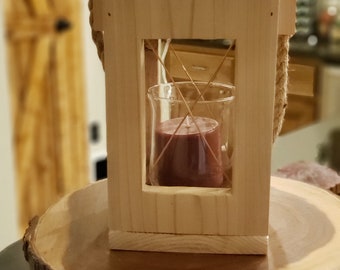 Può includere: Una lanterna per candele in legno con un manico in corda e un portacandele in vetro. La lanterna è appoggiata su una fetta di legno.