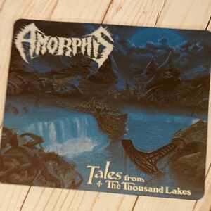 May include: A black and white mousepad with a blue and white image of a lake and mountains. The text "AMORPHIS Tales from The Thousand Lakes" is printed on the mousepad.
