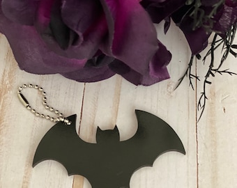 May include: A black bat-shaped keychain with a silver chain. The keychain is on a white wooden surface.