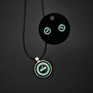 May include: A black necklace with a silver pendant featuring a green circle with a black line through it. The necklace comes with a pair of black earring studs with green circles and black lines.