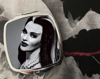 May include: A silver compact mirror featuring a black and white image of a woman with dark hair and dramatic makeup. The image evokes a classic horror film.