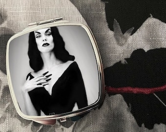 May include: A silver compact mirror featuring a black and white image of a woman with dark hair and dark lipstick. The woman is wearing a black dress with a low neckline.