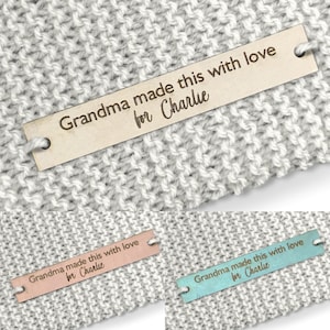Può includere: Un'etichetta di legno con il testo "Grandma made this with love for Charlie" in un font scritto a mano. L'etichetta è attaccata a una coperta di maglia.