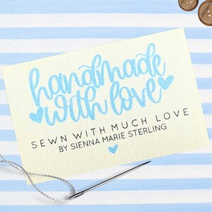 Może przedstawiać: Kremowa metka z napisem "handmade with love" w jasnoniebieskim skrypcie. Poniżej tekst brzmi "SEWN WITH MUCH LOVE BY SIENNA MARIE STERLING". Igła z nitką znajduje się na pierwszym planie, z niebiesko-białym pasiastym tłem.