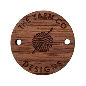 May include: A round wooden tag with the text "The Yarn Co Designs" and a stylized image of a ball of yarn with a knitting needle.