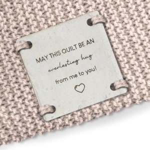 May include: A beige knitted blanket with a white tag attached. The tag has the text "MAY THIS QUILT BE AN everlasting hug from me to you!" with a small heart symbol.