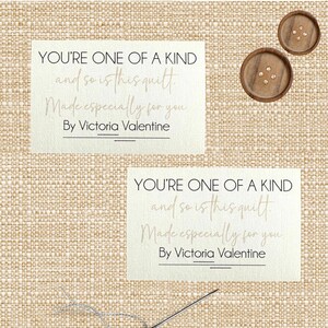 Może przedstawiać: Dwie kremowe karty z tekstem "YOU'RE ONE OF A KIND and so is this quilt. Made especially for you. By Victoria Valentine." Dwa drewniane guziki znajdują się w prawym górnym rogu. Igła i nić znajdują się w dolnej części.