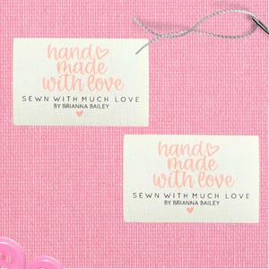 Może przedstawiać: Dwie prostokątne etykiety z napisem "hands made with love" w różowym skrypcie. Poniżej znajduje się tekst "SEWN WITH MUCH LOVE BY BRIANNA BAILEY". Widoczna jest igła i nić. Tło jest różowe.