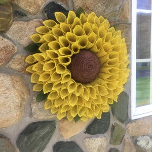 May include: A yellow burlap sunflower wreath with a brown centre. The wreath is hanging on a stone wall.
