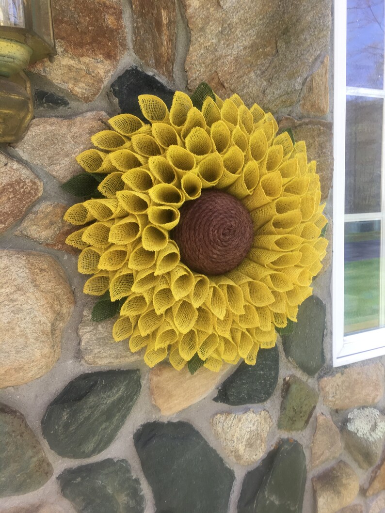 May include: A yellow burlap sunflower wreath with a brown centre. The wreath is hanging on a stone wall.