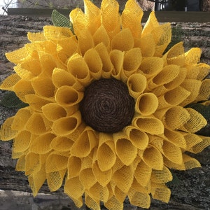 May include: A large, yellow burlap sunflower wreath with a brown burlap centre. The wreath is made of many individual burlap loops that are arranged in a circular pattern.