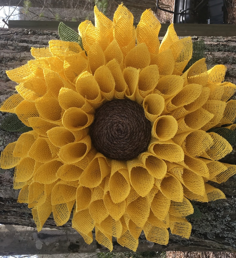 May include: A large, yellow burlap sunflower wreath with a brown burlap centre. The wreath is made of many individual burlap loops that are arranged in a circular pattern.