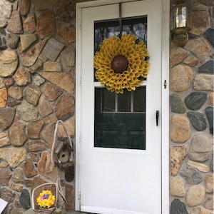 May include: A white front door with a yellow sunflower wreath hanging on it. The door is surrounded by a stone wall. There is a birdhouse and a basket of flowers on the porch.
