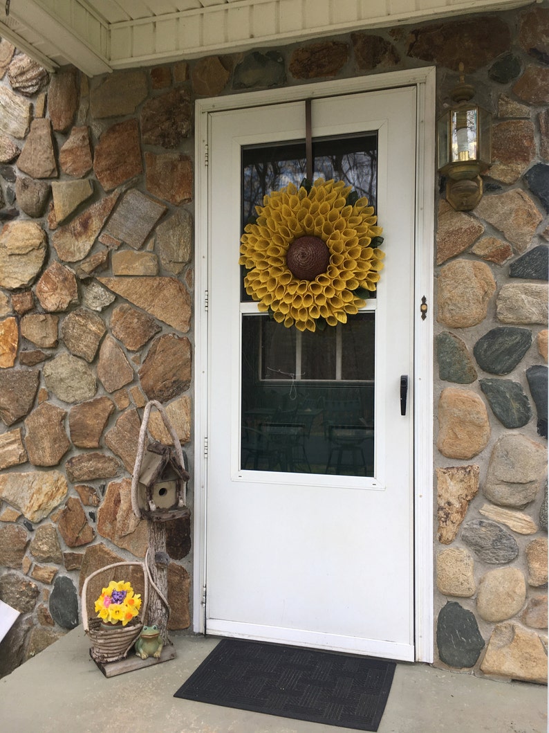May include: A white front door with a yellow sunflower wreath hanging on it. The door is surrounded by a stone wall. There is a birdhouse and a basket of flowers on the porch.