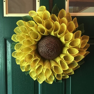 May include: A yellow burlap sunflower wreath with a brown centre. The wreath is hanging on a green door.