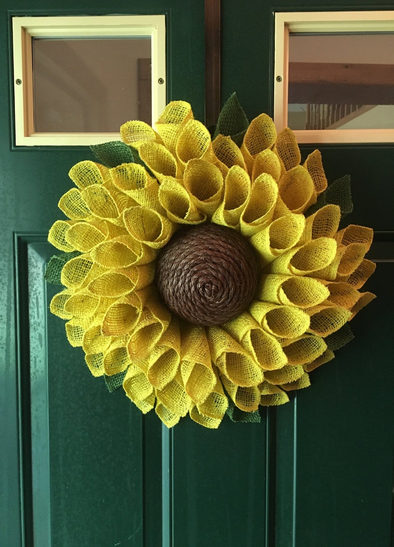 May include: A yellow burlap sunflower wreath with a brown centre. The wreath is hanging on a green door.